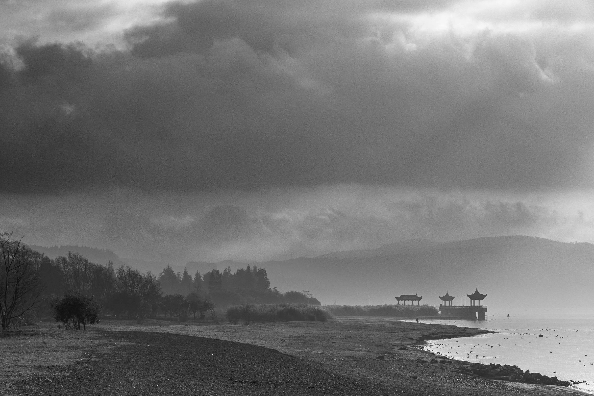 A black-and-white lakeshore beneath layered storm clouds, with distant hills and pavilion silhouettes beside the water.