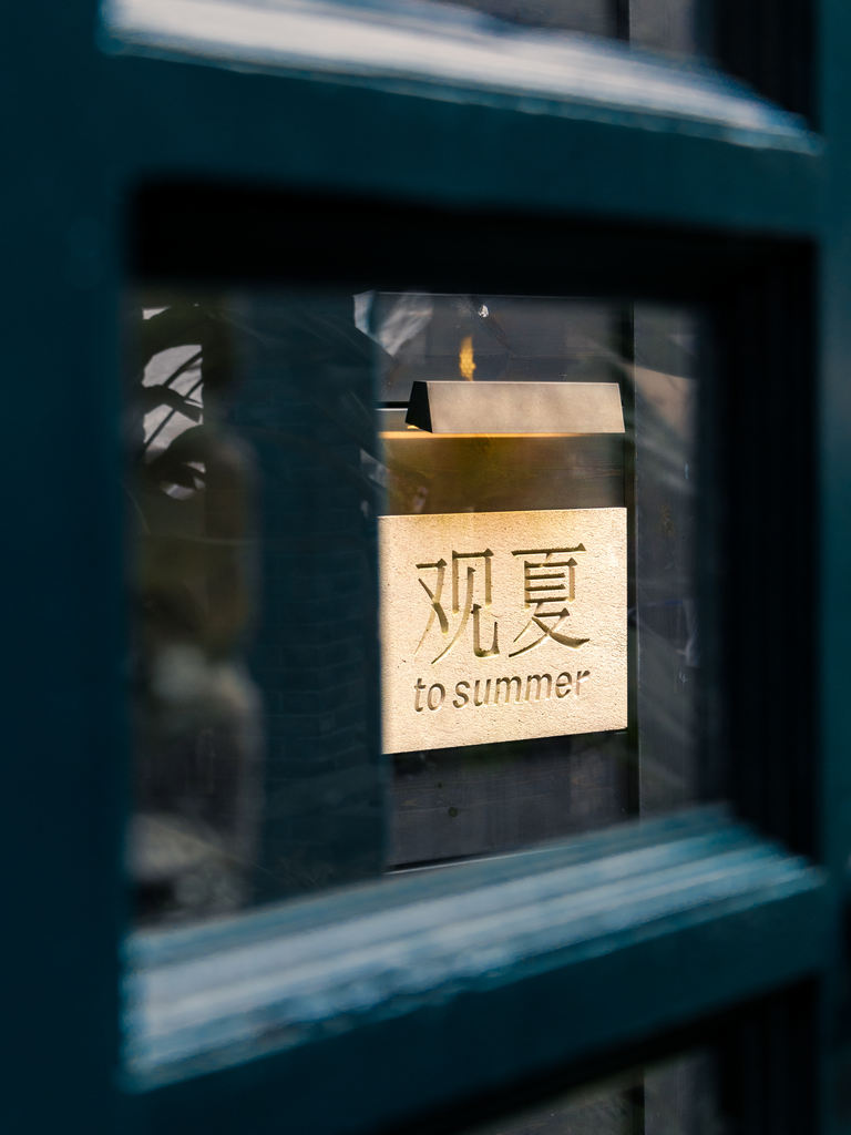 A glowing 观夏 / To Summer sign seen through dark blue storefront framing.